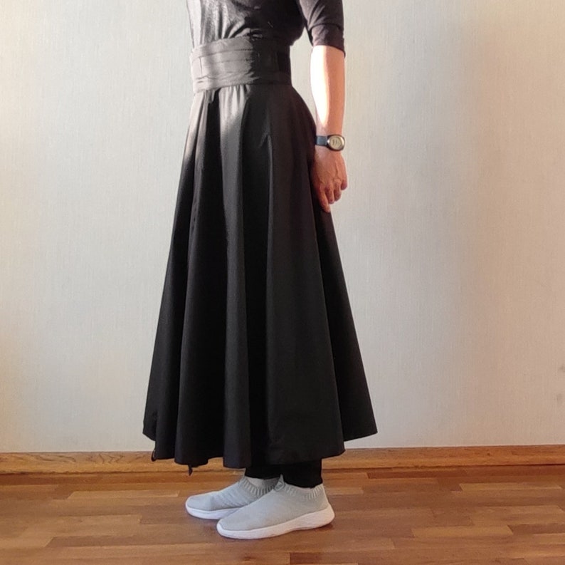 May include: A black, full-length skirt with a wide waistband. The skirt is paired with a black top and light gray slip-on sneakers. A watch is visible on the wrist. The skirt has a flared silhouette.
