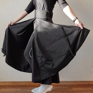 May include: A black wrap skirt with a wide waistband and a flowing, asymmetrical design. The skirt is made of a lightweight fabric and features a belt closure. The model is wearing a black top and gray leggings.