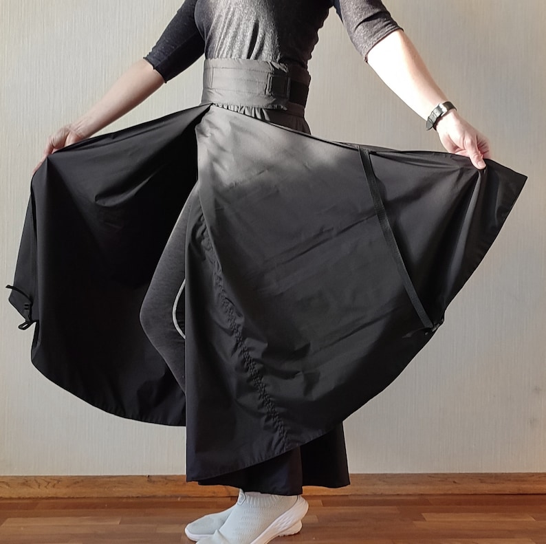 May include: A black wrap skirt with a wide waistband and a flowing, asymmetrical design. The skirt is made of a lightweight fabric and features a belt closure. The model is wearing a black top and gray leggings.