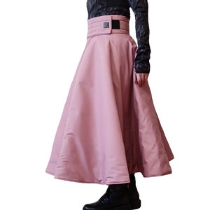 May include: A full-length, light pink skirt with a wide, quilted waistband. The skirt has a flared silhouette and a smooth, matte finish. The waistband features a black rectangular detail. The model is wearing black boots and a black top.