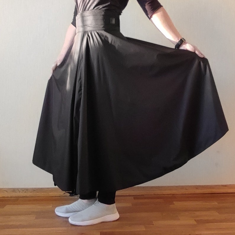 May include: A black skirt with a wide waistband and a flowing, full silhouette. The skirt is made of a smooth, dark fabric and is being held up to show its shape. The person is wearing black leggings and gray slip-on sneakers.