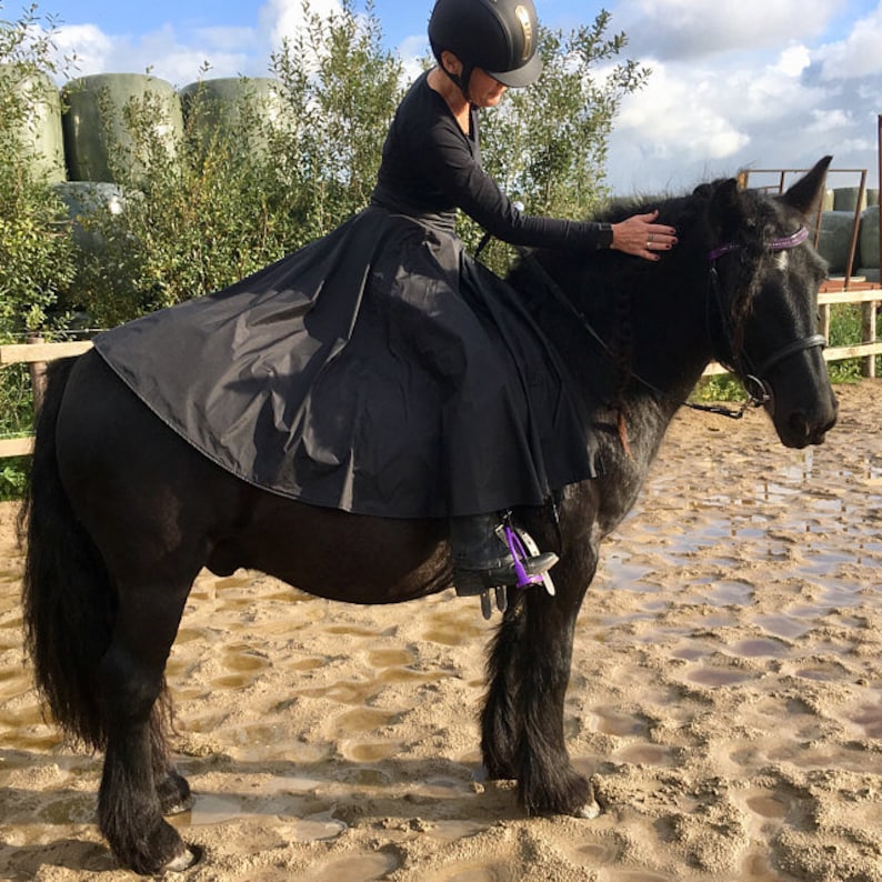 May include: A black horse with a rider wearing a black riding skirt and helmet. The horse has a black mane and tail, and is standing in a sandy area. The rider is petting the horse.