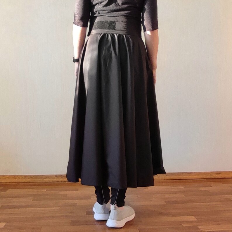 May include: A black, full-length skirt with a wide waistband and a flared silhouette. The skirt is paired with black leggings and light gray sneakers. The person is standing in front of a light-colored wall and a wooden floor.