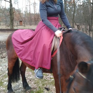May include: A person riding a brown horse outdoors, wearing a dark top and a long, burgundy skirt. The horse has a dark mane and tail. The scene is set in a wooded area with trees in the background.