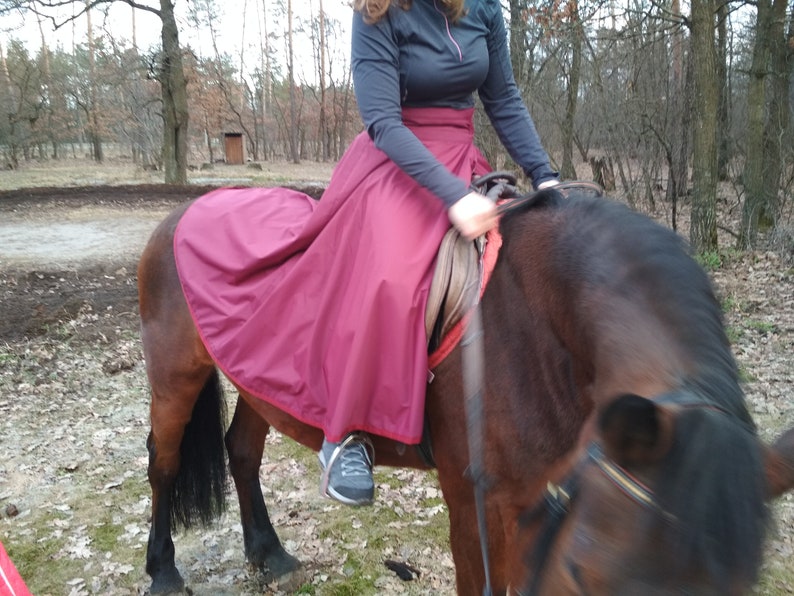 May include: A person riding a brown horse outdoors, wearing a dark top and a long, burgundy skirt. The horse has a dark mane and tail. The scene is set in a wooded area with trees in the background.