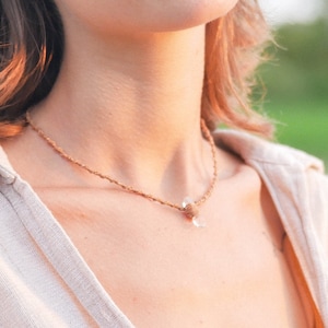 Puede incluir: Una mujer con el pelo largo y castaño lleva una camisa beige claro con un collar de cordón marrón con un pequeño colgante de cristal.