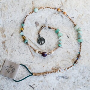May include: A brown cord necklace with green, brown, and clear beads, a purple stone, and a tree of life charm. The necklace is coiled on a white textured surface.