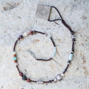 May include: A brown cord necklace with various coloured beads, including brown, red, pink, green, and clear. A silver feather charm hangs from the necklace.