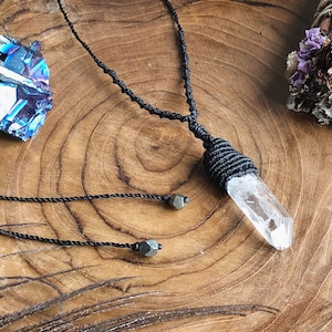 May include: A clear quartz crystal pendant on a black macrame cord necklace with two black hematite beads. The necklace is displayed on a wooden surface.