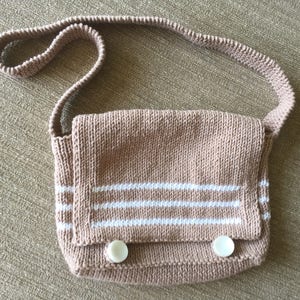 Könnte beinhalten: Eine braune gestrickte Umhängetasche mit weißen Streifen und zwei weißen Knöpfen. Die Tasche hat einen langen Riemen.