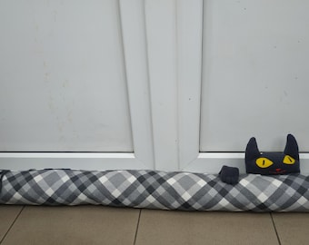 Può includere: Un paraspifferi cilindrico e lungo in tessuto con un motivo a quadri grigio, bianco e nero. Sulla parte superiore è fissata una testa di gatto in peluche nera con occhi gialli e piccole orecchie, insieme a due piccole zampe nere sul tessuto.
