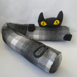 以下が含まれることがあります： 黄色い目をした黒と白のチェック柄の猫の形をしたドアストッパー。猫は横になっていて、頭と足が突き出ています。
