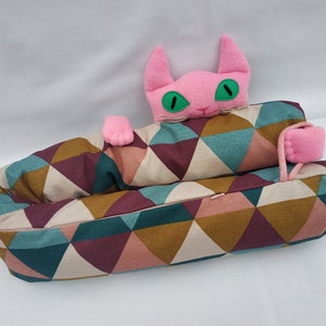 Peut inclure: Une tête de chat rose émerge d'un long coussin cylindrique avec un motif géométrique de triangles bruns, roses, turquoise et blancs.