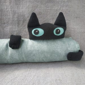 May include: A plush black cat head with large teal eyes attached to a long, cylindrical, light green bolster pillow. The cat has two black paws extending from the sides of the pillow, which features a textured fabric surface.