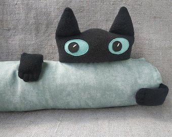 May include: A plush black cat head with large teal eyes attached to a long, cylindrical, light green bolster pillow. The cat has two black paws extending from the sides of the pillow, which features a textured fabric surface.
