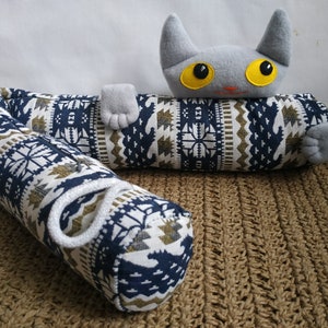 Puede incluir: Un juguete de peluche con forma de gato gris con ojos amarillos está tumbado sobre una almohada cilíndrica con estampado azul y blanco. La almohada tiene una cuerda blanca atada alrededor.