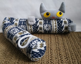 Può includere: Un giocattolo di peluche a forma di gatto grigio con occhi gialli è sdraiato su un cuscino cilindrico con motivo blu e bianco. Il cuscino ha una corda bianca legata intorno.