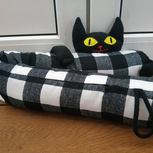May include: Black and white checked doorstop shaped like a cat with yellow eyes. The cat is lying down with its head and paws sticking out.