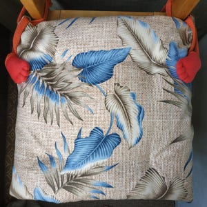 Puede incluir: Cojín de silla con un estampado de hojas tropicales en tonos azules, grises y beige. El cojín tiene un aspecto texturizado y está adornado con cuerda naranja y detalles rojos.