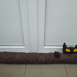 May include: A brown fabric doorstop shaped like a cat with yellow eyes. The doorstop is lying on a tiled floor.