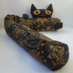 Può includere: Un fermaporta nero e dorato a forma di bastone di zucchero con una testa di gatto nero in cima. Il gatto ha occhi gialli e un naso rosso. Il fermaporta è rivestito di un tessuto nero con un motivo a fiocco di neve dorato.