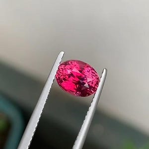May include: A faceted, oval-shaped, vibrant red gemstone is held by silver tweezers. The gem's color is a deep, saturated pink-red, with visible internal reflections. The tweezers are in sharp focus, highlighting the gem's clarity.