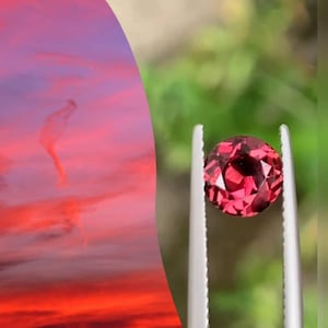 以下が含まれることがあります： ファセット加工された丸い深紅色の宝石が、ぼやけた緑色の背景に対して銀色のピンセットで保持されています。宝石の色は、夕焼け空の鮮やかな色合いに似ており、赤と紫の色合いがあります。