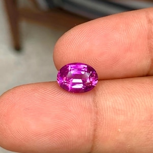 May include: A vibrant, oval-cut, magenta gemstone. The gem is held between fingers, showcasing its facets and internal structure. The stone's color is a rich, saturated pink, reflecting light and creating a sparkling effect. This gemstone is suitable for jewelry.