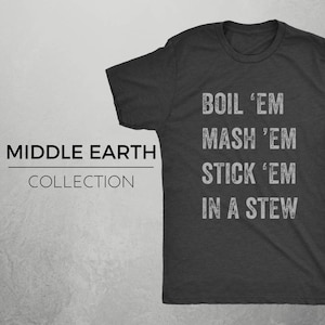 Op de afbeelding: Donkergrijs T-shirt met witte tekst die "BOIL 'EM MASH 'EM STICK 'EM IN A STEW" luidt. De tekst is in een verweerd lettertype. Het shirt maakt deel uit van een collectie genaamd "MIDDLE EARTH COLLECTION".