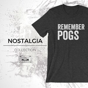 May include: A dark heather grey t-shirt with the text "Remember POGS" in white lettering. The shirt is part of a "Nostalgia Collection" and features a vintage cassette tape graphic.