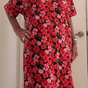 Op de afbeelding: Een jurk met korte mouwen en knielengte, voorzien van een levendige all-over bloemenprint met rode, roze en lichtroze hibiscusbloemen op een zwarte achtergrond. Het kledingstuk heeft een eenvoudige ronde hals met een effen rode bies.