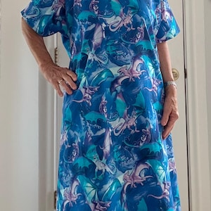 Op de afbeelding: Een jurk met korte mouwen en knielengte, voorzien van een all-over print van draken in tinten blauw, taling en lichtpaars. Het kledingstuk heeft een ronde halslijn met een donkerblauwe bies en een losse, recht gesneden pasvorm.