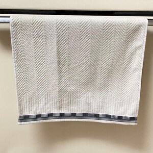 Op de afbeelding: Een witte keukendoek met een herringbone-patroon en een zwart-groene gestreepte rand die aan een zilveren handdoekrek hangt.
