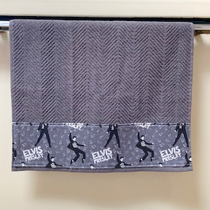 Op de afbeelding: Een donkergrijze handdoek met een gestructureerd visgraatpatroon op het bovenste gedeelte en een decoratieve stoffen band aan de onderkant. De band toont een herhalende print van Elvis Presley in een danshouding met de tekst Elvis Presley.
