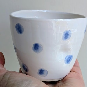 Könnte beinhalten: Ein weißer Keramikbecher mit glänzender Oberfläche, verziert mit einem handgemalten Muster aus verstreuten blauen Punkten. Der Becher hat eine leicht unregelmäßige, abgerundete Form mit einer weiten Öffnung und einer glatten, gewölbten Außenseite.