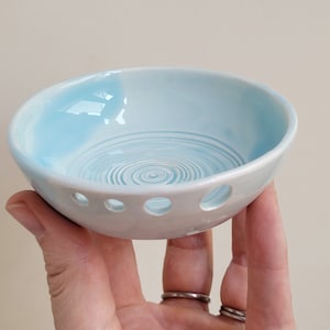 May include: A light blue ceramic bowl with a white interior and a swirling pattern. The bowl has drainage holes around the bottom.