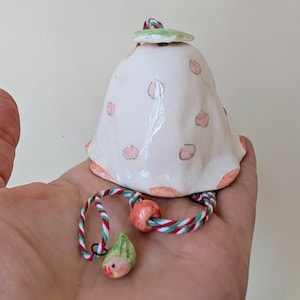 May include: A white ceramic bell with a glossy finish and hand-painted peach-coloured polka dots. The bell features a green leaf-shaped top and a hanging cord made of red, white, and green twisted string with a small peach bead and a green charm.