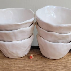 May include: Six white ceramic bowls with a textured, organic design, arranged in pairs. They sit on a wooden surface. A small bird figurine is placed in front of the bowls. Each bowl has a unique shape.