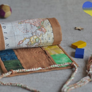 May include: A brown fabric pencil roll with a world map design and a tassel. The roll is unrolled and shows a brown fabric base with colourful fabric strips. The roll is on a light grey surface with two crayons, two wooden blocks, and a wooden star.