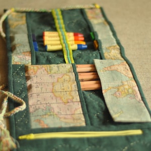 May include: A green fabric pencil roll with a map print. The roll has a zipper closure and a tassel. The roll is filled with coloured pencils and crayons.