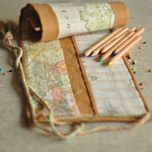 Puede incluir: Un estuche enrollable de tela marrón con estampado de mapa y un borla. El estuche está abierto y tiene una fila de lápices de colores dentro.