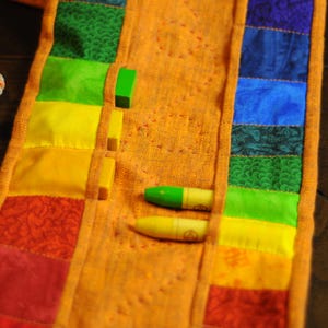 May include: A colorful fabric strip with pockets for crayons. The fabric is orange with rainbow colored stripes and a floral pattern. Two crayons are visible in the pockets.