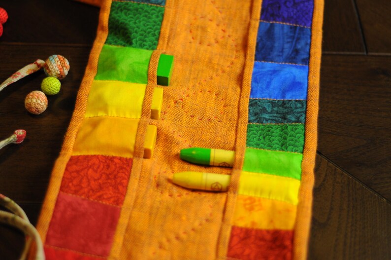 May include: A colorful fabric strip with pockets for crayons. The fabric is orange with rainbow colored stripes and a floral pattern. Two crayons are visible in the pockets.