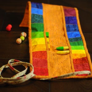 May include: A rainbow fabric roll-up case with multiple pockets for storing crayons or colored pencils. The case is made of a patchwork of colorful fabrics and has a brown fabric strap.