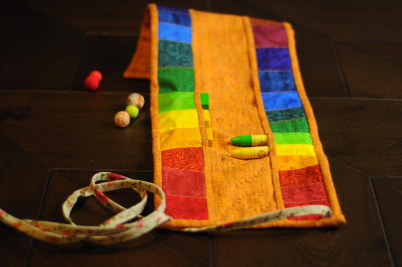May include: A rainbow fabric roll-up case with multiple pockets for storing crayons or colored pencils. The case is made of a patchwork of colorful fabrics and has a brown fabric strap.