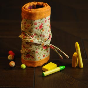 May include: A floral fabric pencil roll with a yellow and orange trim. The roll is unrolled and there are several yellow and orange crayons and a green eraser on the table.