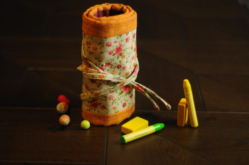 May include: A floral fabric pencil roll with a yellow and orange trim. The roll is unrolled and there are several yellow and orange crayons and a green eraser on the table.