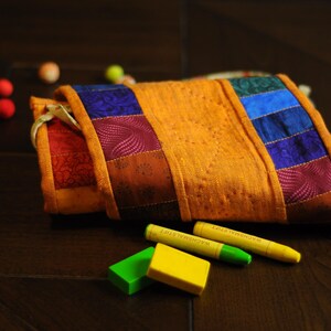 May include: A colorful patchwork fabric pouch with a yellow, orange, red, blue, and purple design. The pouch is lying on a dark wood surface with two green and yellow crayons next to it.