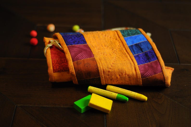 May include: A colorful patchwork fabric pouch with a yellow, orange, red, blue, and purple design. The pouch is lying on a dark wood surface with two green and yellow crayons next to it.