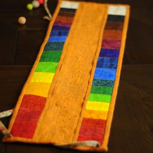 May include: A long, orange fabric pouch with a rainbow of colors sewn along the edge. The pouch is folded in half and has a white string tie.